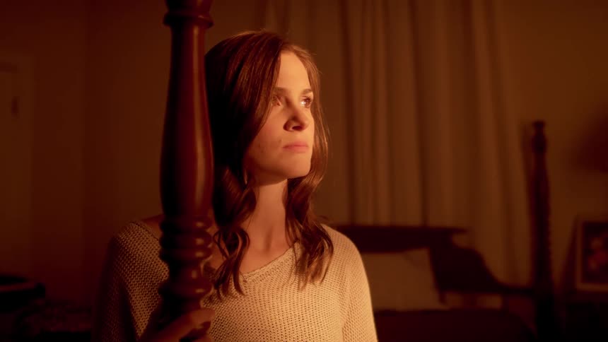 A woman holds onto a bedpost as she calmly looks upwards at a source of light