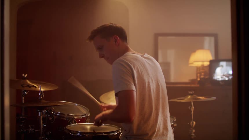 A young male sits and plays a drum set inside an apartment