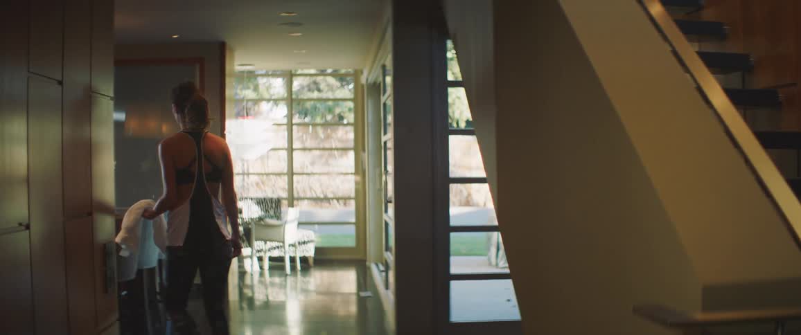 Woman wearing sports bra walks past staircase along hallway toward another room