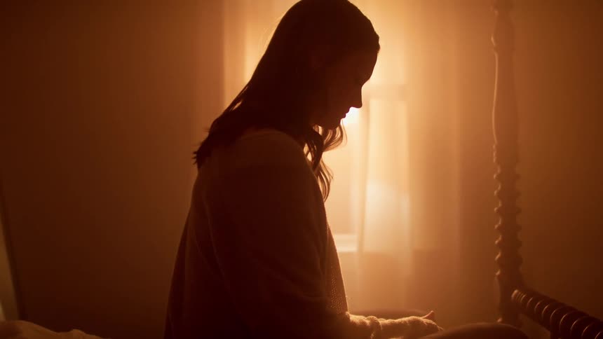 A teenage girl sits on her bed and looks down at her lap