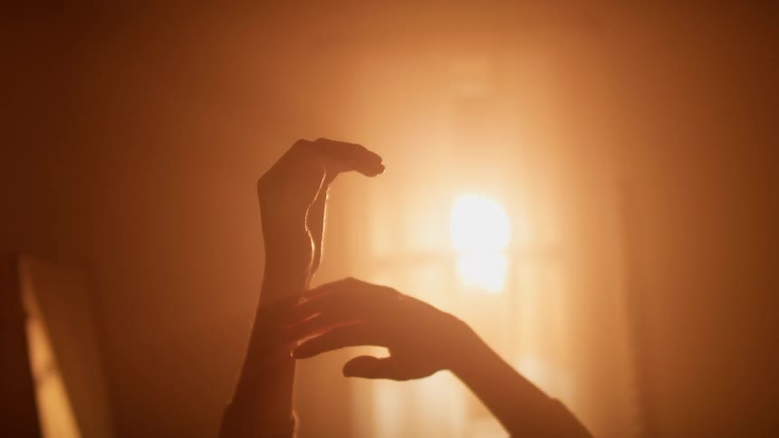 A woman lies in bed and stretches her arms up as sunlight shines through her window and turns the bedroom orange