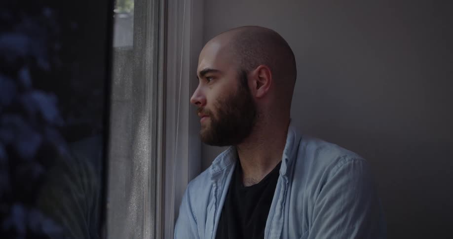 Bald man with a beard and a blue shirt looks out the window