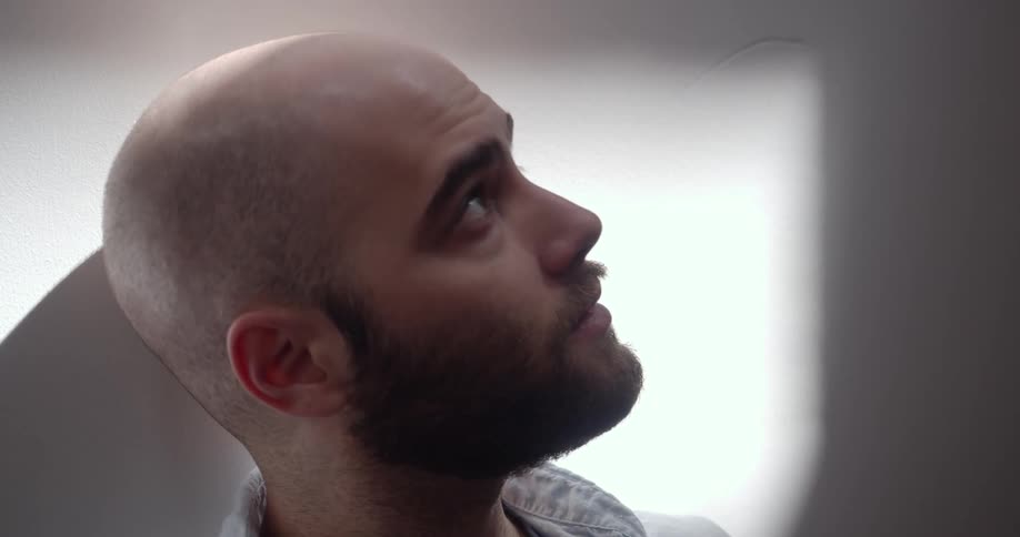 Bald man with a beard sits down and leans against the wall in a room