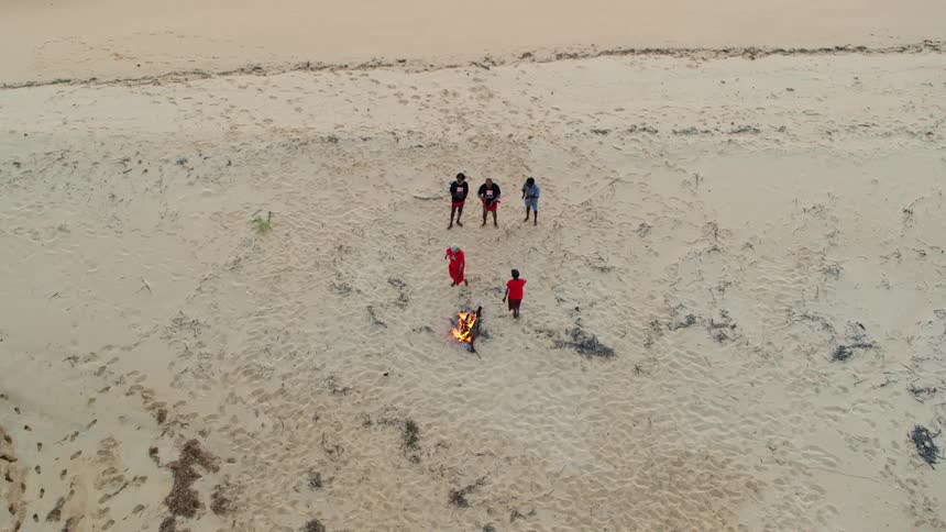 People dance on sand around small bonfire as others clap