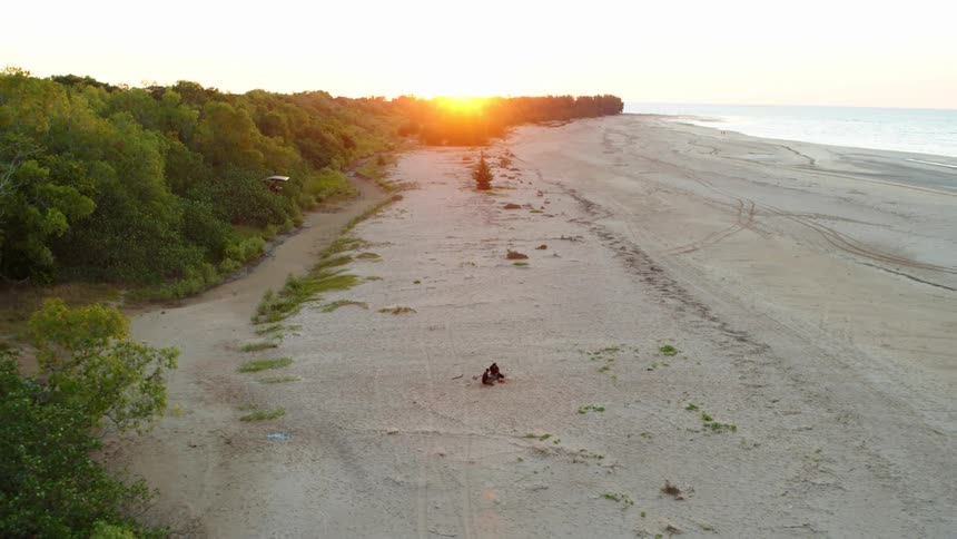 Two people sit on the sand of an empty beach as the golden sun shines on the trees