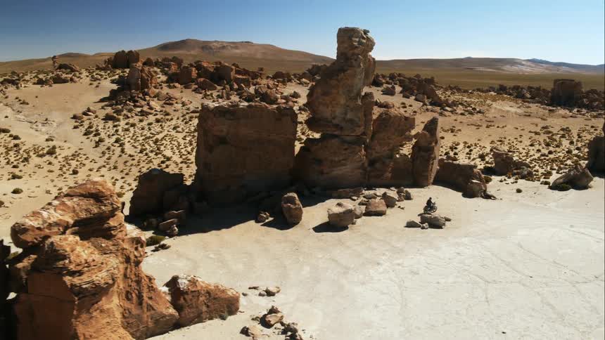 A biker rides a motorcycle offroad past rock formations in the desert during the day