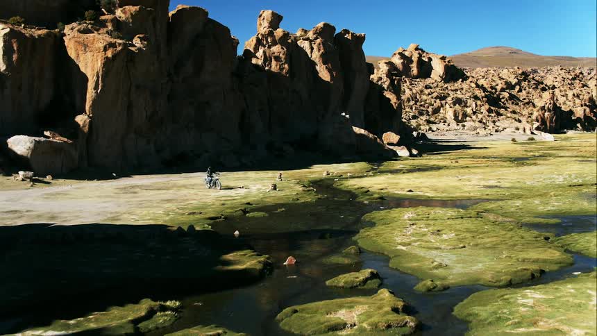 A person on a motorcycle drives past cliffs and up to a river bed