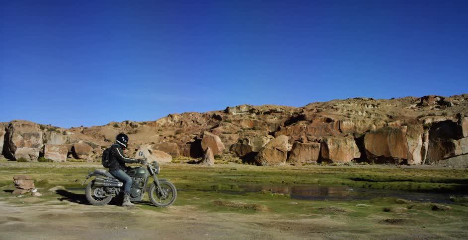 A person with a backpack and a helmet on a motorcycle with a cliff the sky and clouds in the background