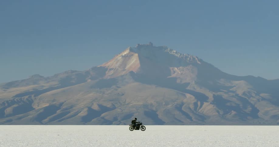 Man rides a motorcycle through the desert with a mountain in the background