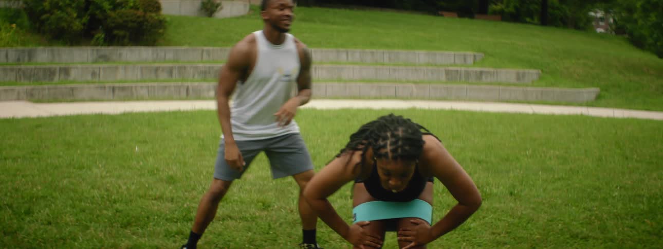 A woman with a resistance band around her thighs exercises beside a man in a field in a park during the day