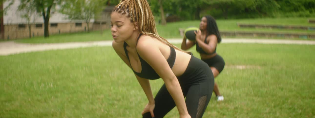 A woman takes a break from working out in a park while a woman behind her lifts and squats with a medicine ball
