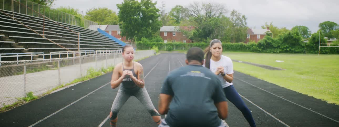 A man leads a pair of young women in exercising on an athletics track before the group bursts into laughter