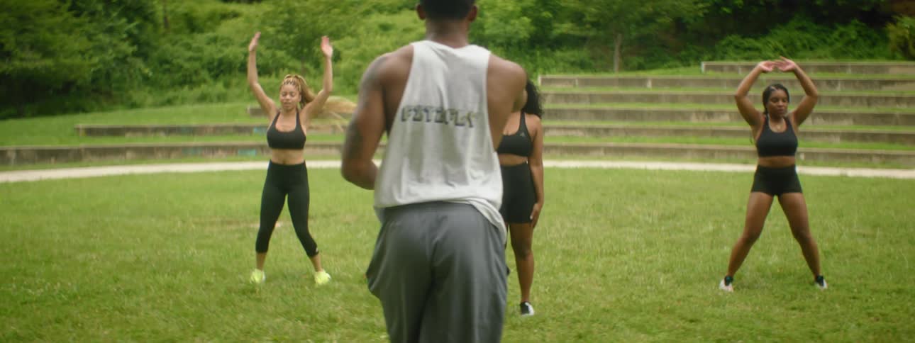 A man leads a trio of women in jumping jack exercises in a park before they all stop and rest