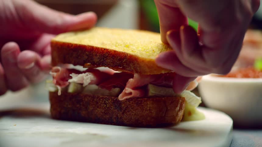 A cook places a toasted slice of bread with sauce on it atop a sandwich on a cutting board