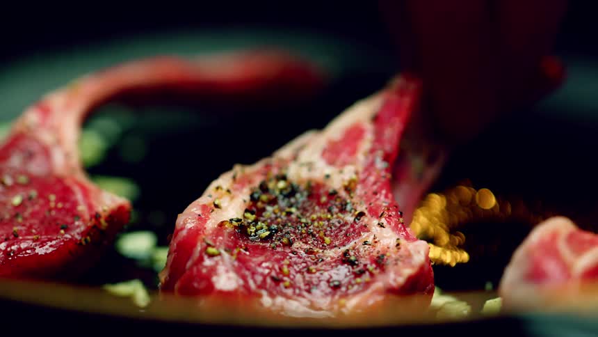 A seasoned piece of red meat is being gently laid into a cast iron skillet containing other pieces of red meat