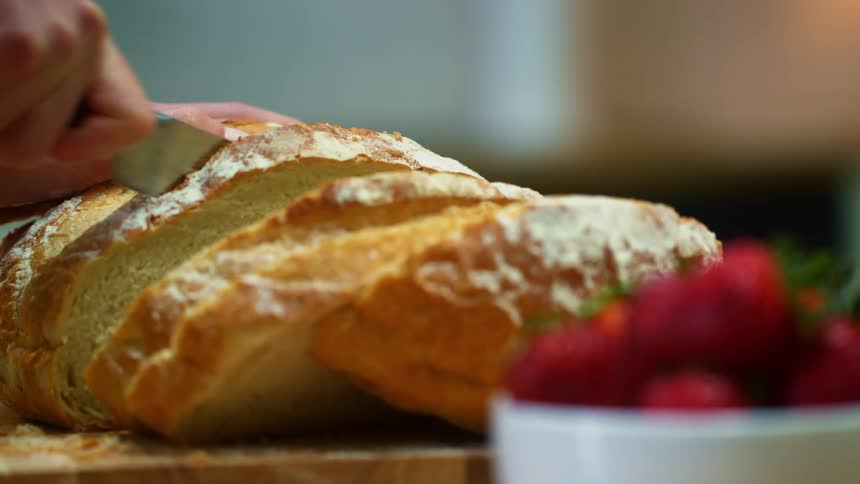 A person uses a bread knife to slice a loaf of bread on a wooden cutting board