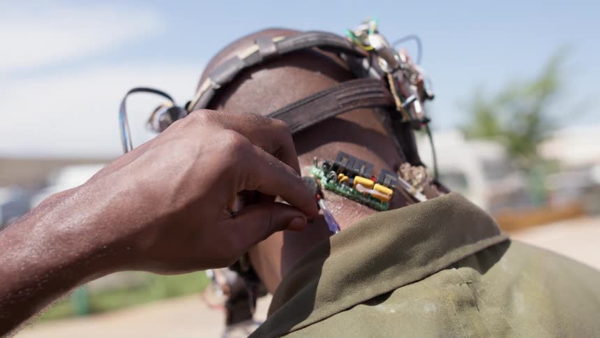 Bald man wearing a futuristic makeshift headset plugs a cable to the implants on the base of his neck