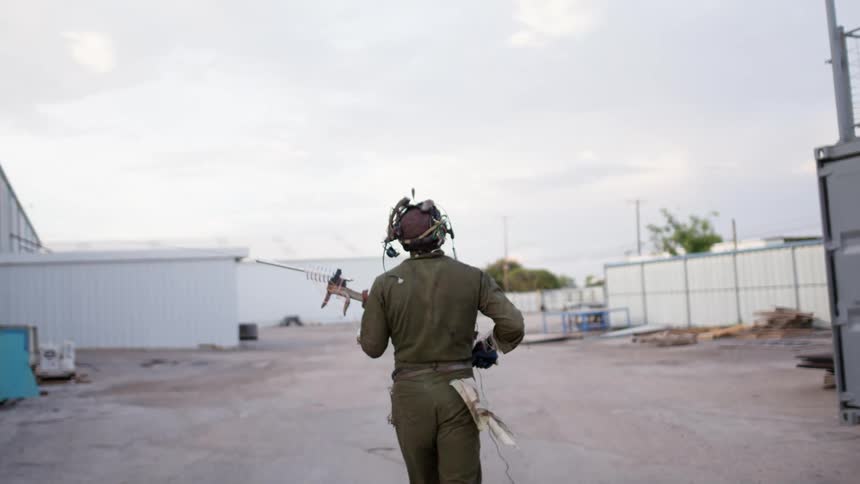 A man walks through a lot with a helmet on with wires as he carries a futuristic looking gun