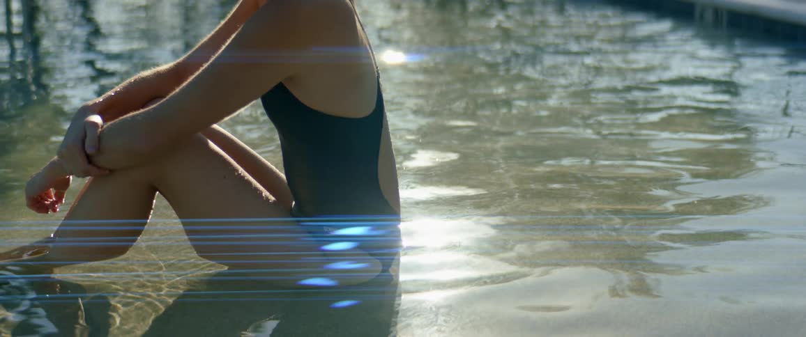 A woman wearing a one piece swimsuit sits outside on a sunny day in a very shallow pool of water