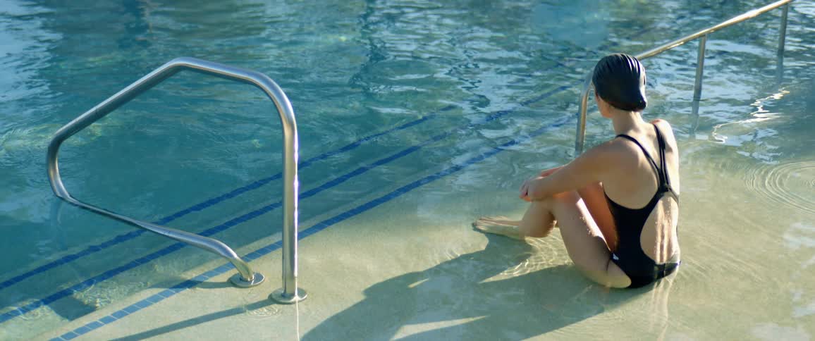 Woman in swimsuit and swim cap sits at edge of swimming pool