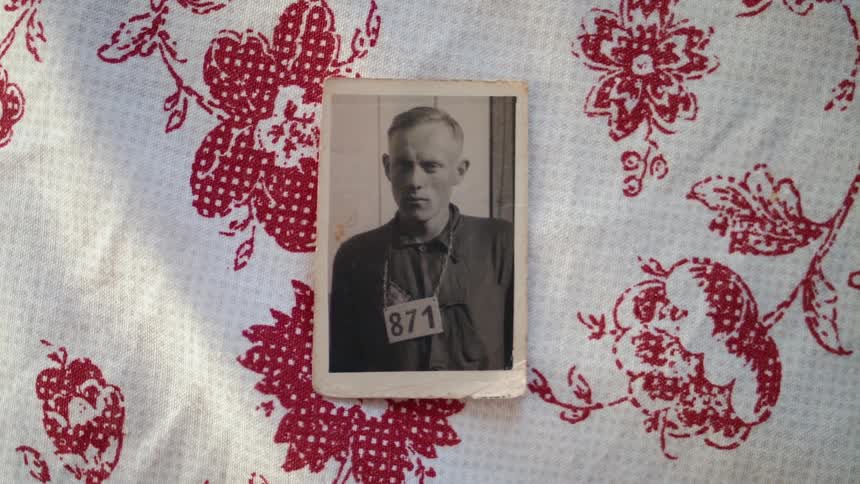 A black and white photo of a man with a three digit number hanging from his neck is taped to a red and white flower print fabric