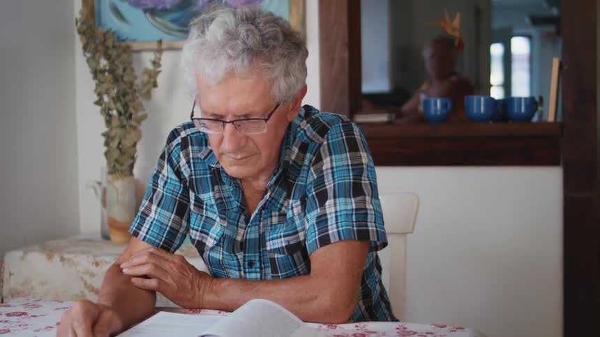A man reads a book at his dining room table while his wife cleans in the kitchen