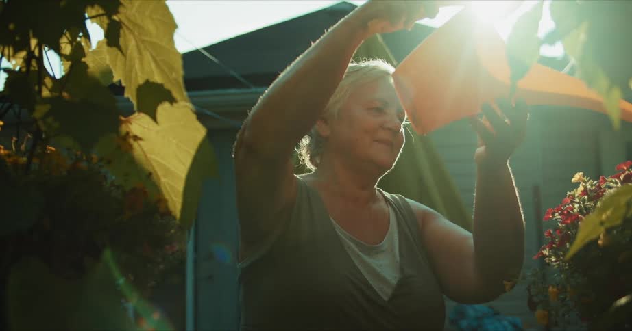 An old caucasian woman with white hair holding a yellow water pitcher is standing outside watering plants on a sunny day