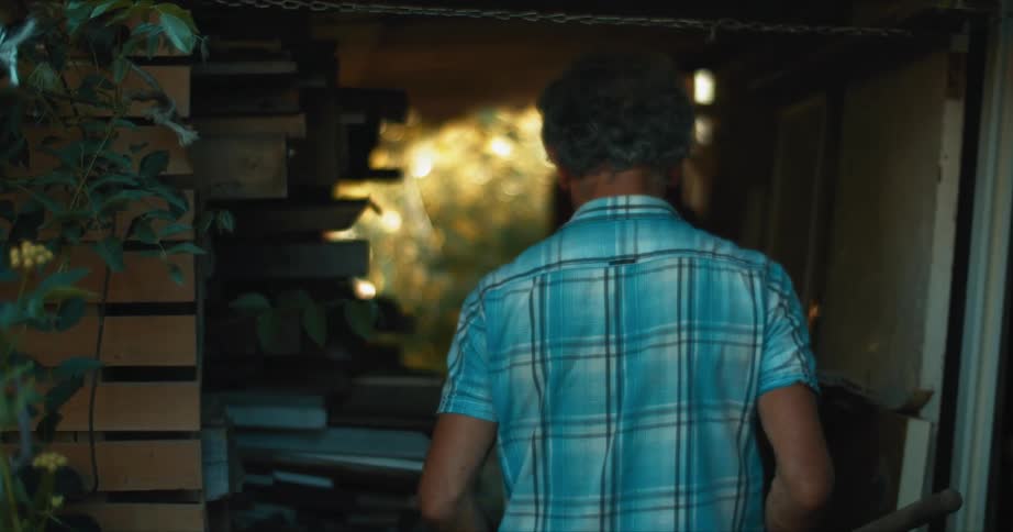 A man carries a shovel in his right hand as he walks past tall plants and enters his shed