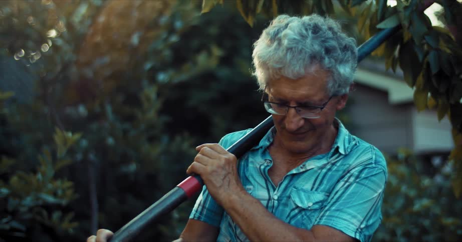 An elderly man in his yard removes a shovel from over his shoulders