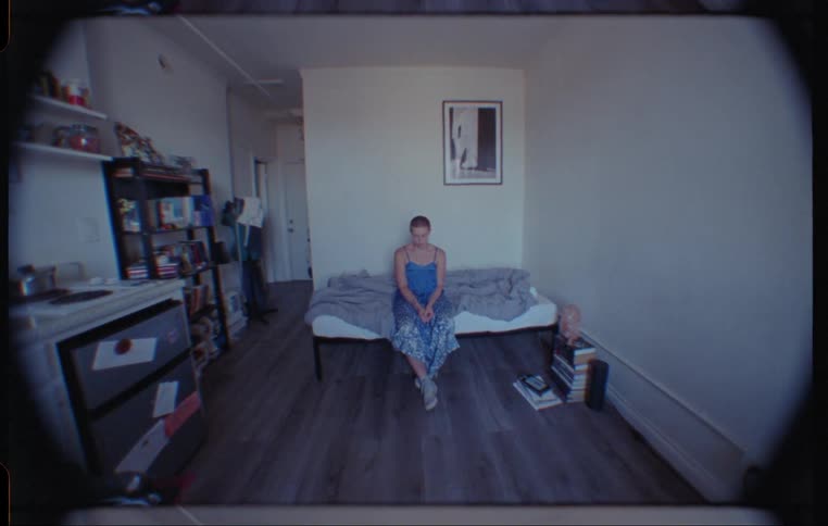 A woman sits quietly on a small apartment bed appearing depressed