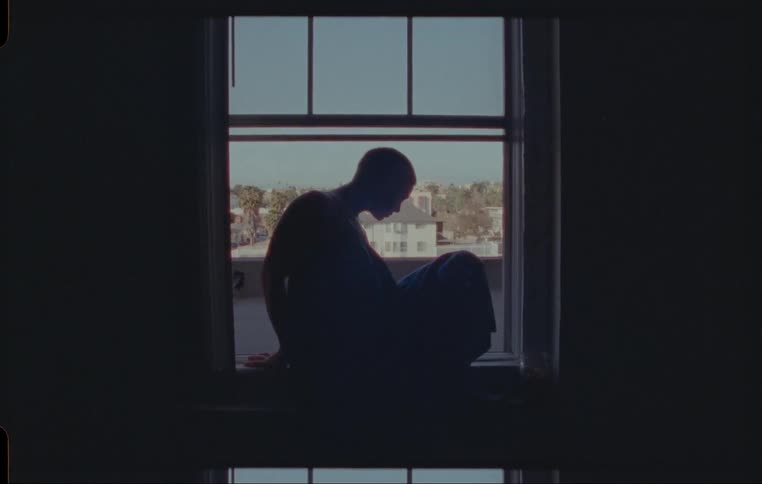 A woman in a dark room approaches and climbs out of a window onto the roof of a building
