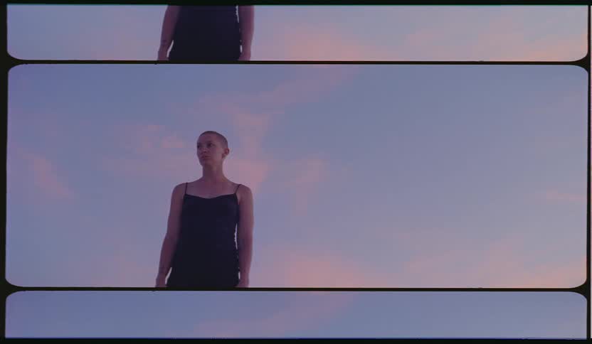 A woman with a shaved head stands outside in front of a pink and blue sky