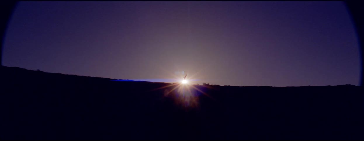 A person is dancing and running back and forth on a hilltop in silhouette with the sun glaring behind them