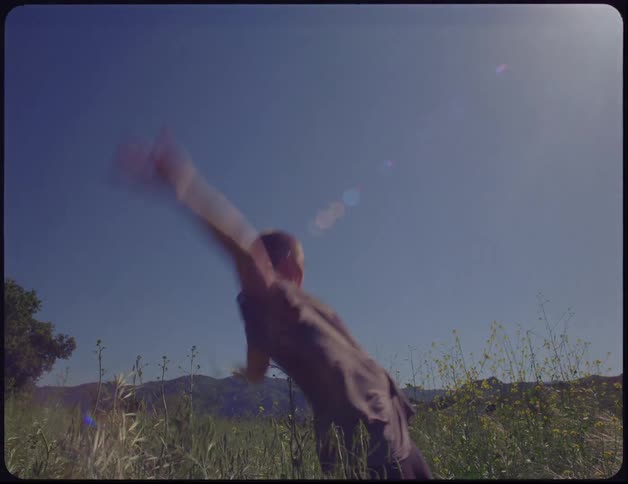 Woman with a buzz cut and a purple dress dances in a grass field during the day