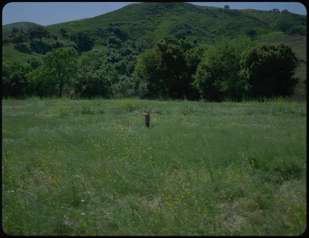A woman dances in a grassland below a tall hill on a sunny day