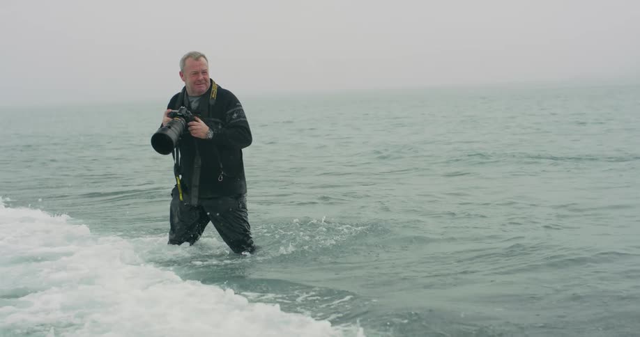 Ocean waves crash against a photographer as he stands in the ocean in an attempt to take a photograph