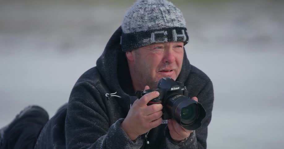 A man lies down on the ground and raises his camera to his eye to take a photo
