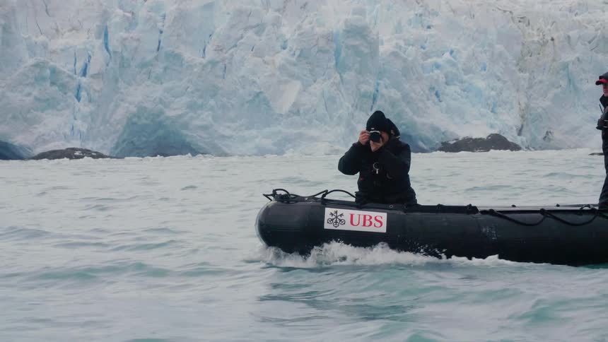 A man with a camera a coat and a hat on a boat with glaciers and rocks in the background