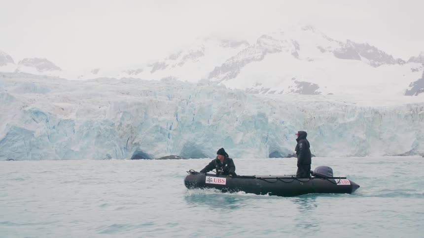 A man traveling on a motorboat in the arctic with another man turns toward the camera and gives a thumbs up