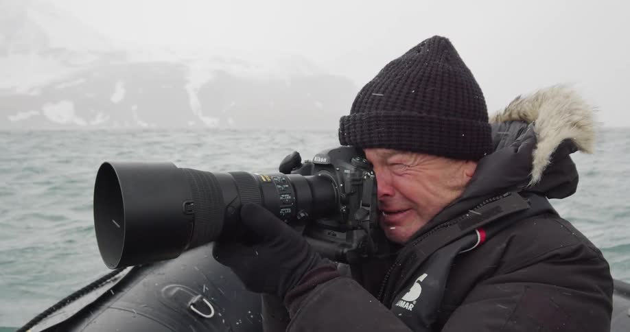 Photographer bobs in small boat using telephoto lens while snow and fog obscure mountain slope on horizon