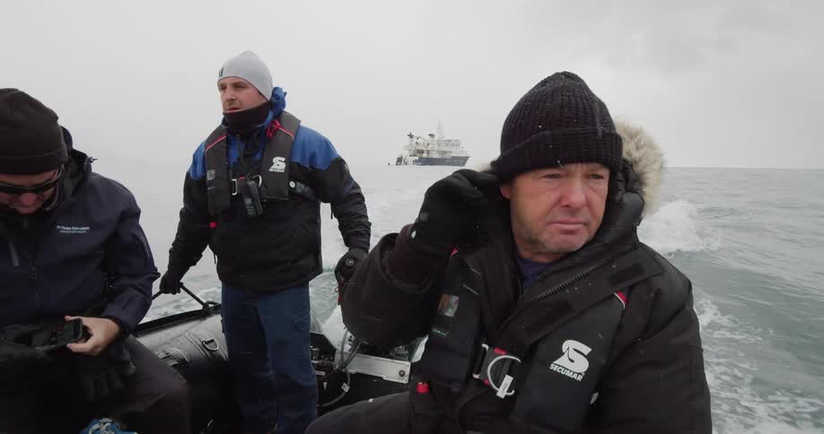 A group of men in winter clothing ride a raft across the ocean away from a ship as snow falls from a cloudy sky