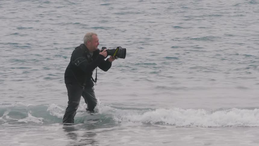 A photographer stands in the water on the shore as he takes photos