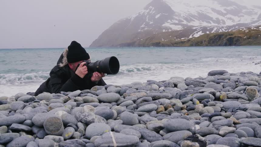 A photographer leans against rocks to take photos as a penguin frolics in the ocean behind him