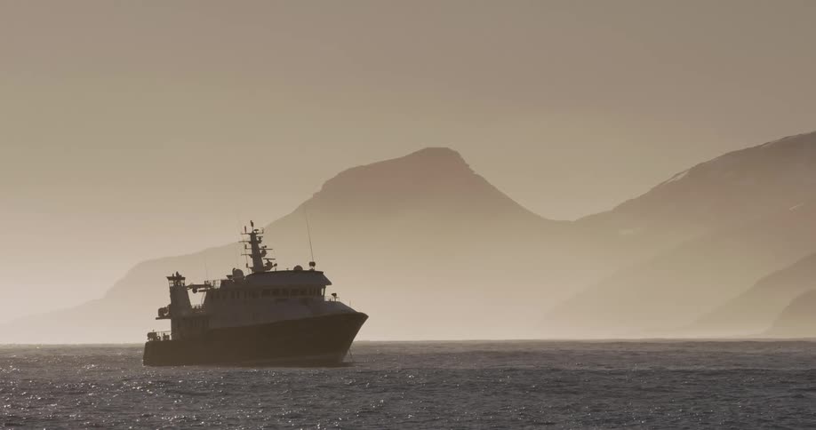 A large boat with lots of instrumentation and antennas on top of it floats near a mountainous shoreline