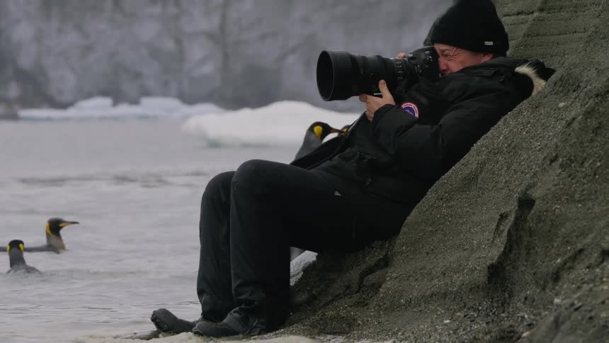 A male photographer sits against a rock taking photos of penguins in arctic water