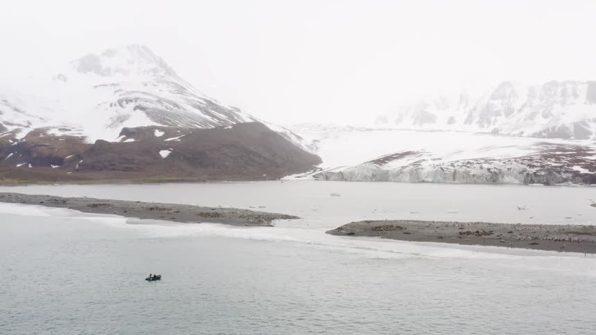 A raft travels towards a gap between a pair of sand bars on the ocean just of the coast of a snowy mountain ridge