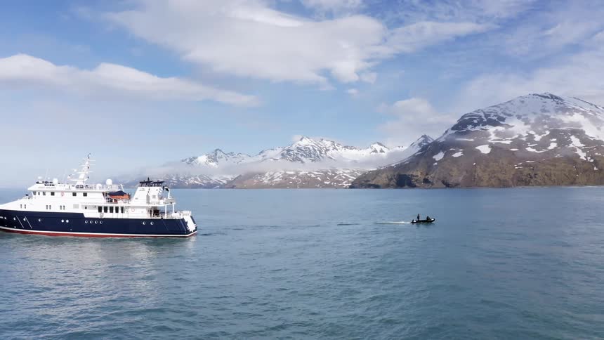 Man stands in small boat as cruise ship sails in the opposite direction both paralleling snow covered mountains sitting on shore under a cloudy sky