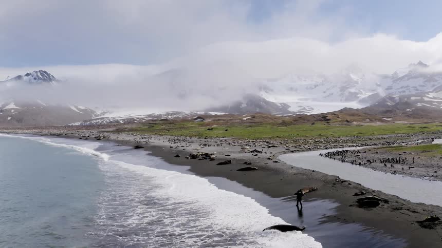 A person walks down a rocky beach with mountains in the distance at midday