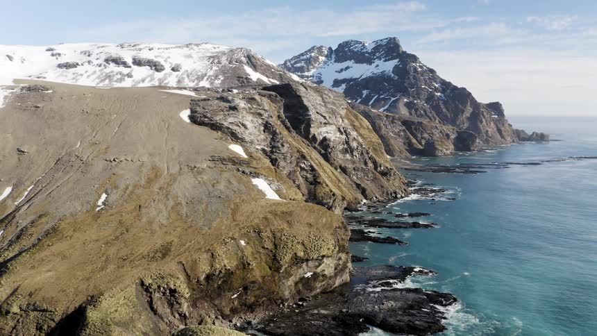 Snow dots slopes of mountains sitting on sea cost as waves crash against the rocks