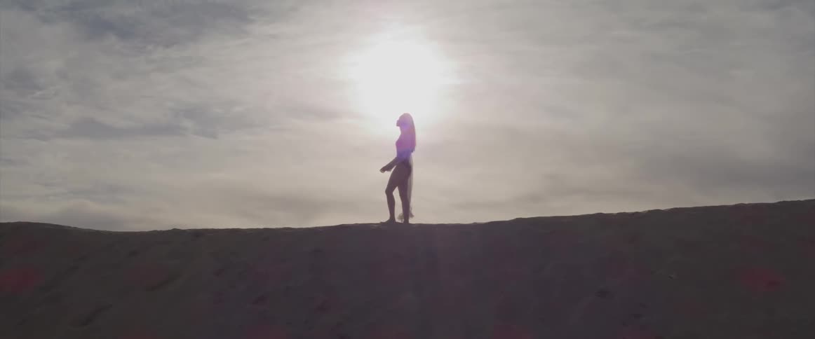 Woman wearing swimsuit walks across top of sandy hill under bright sun shining from cloudy sky